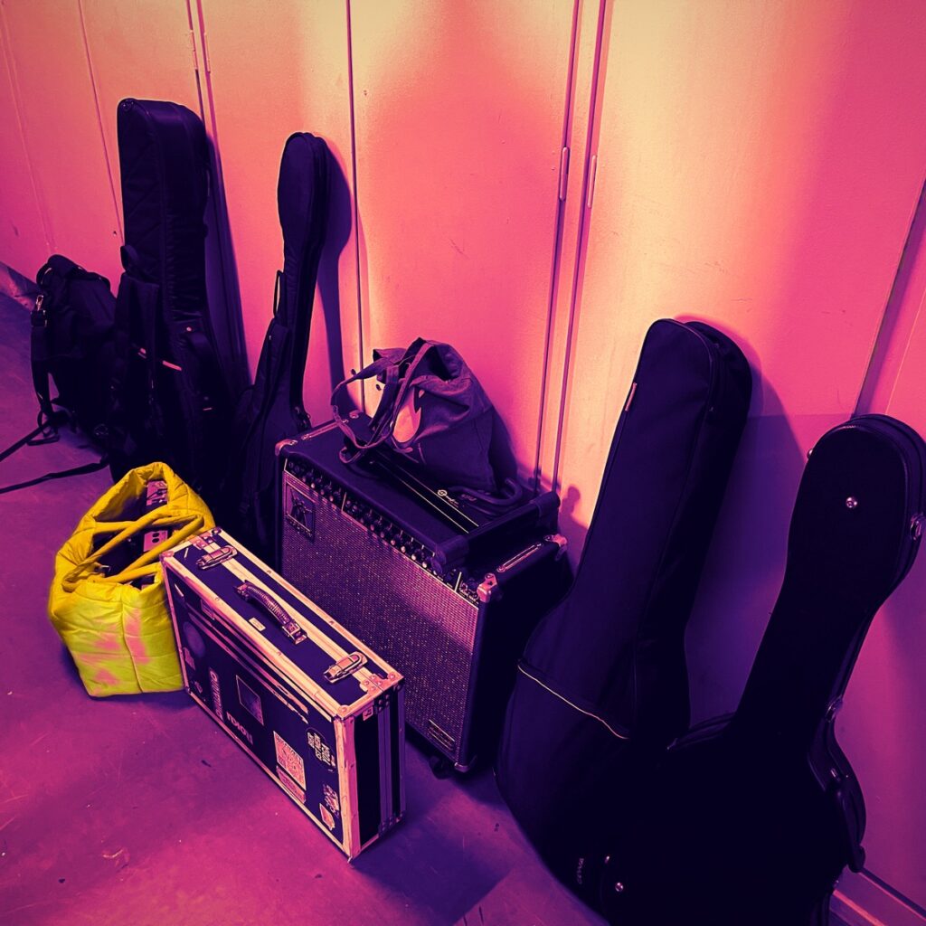 Guitar cases, an amp and a flight case lined up against a wall like luggage, lit in pink and violet